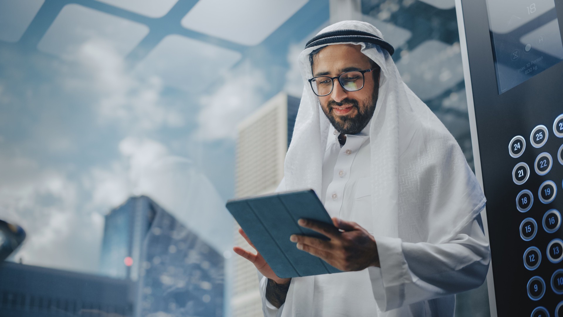 Successful Muslim Businessman in Traditional White Kandura Riding Glass Elevator to Office in Modern Business Center. Man Using Tablet Computer. Saudi, Emirati, Arab Businessman Concept.